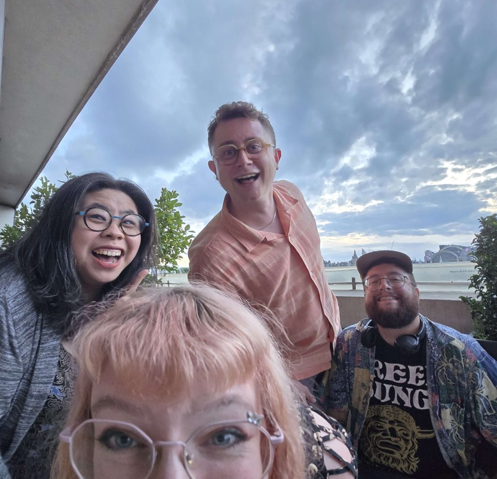 Krya, Jade, Ed and Harvey stand in staggered poses smiling at the camera with a cloudy skyline behind them